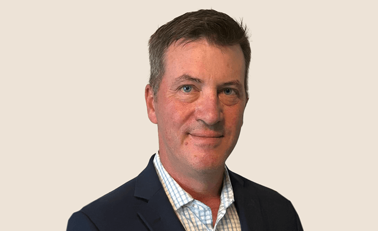 A professional headshot of a middle-aged man smiling, wearing a dark blue suit jacket over a light checkered shirt.
