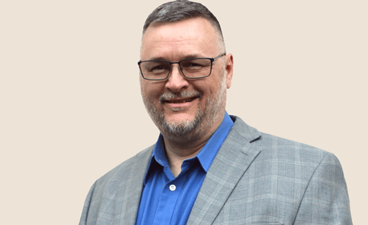 A professional headshot of a smiling man with a beard and glasses, wearing a blue shirt and a gray plaid jacket
