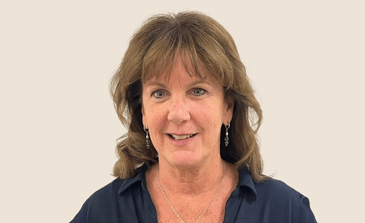 A professional headshot of a smiling middle-aged woman with brown hair and wearing a dark collared shirt and earrings