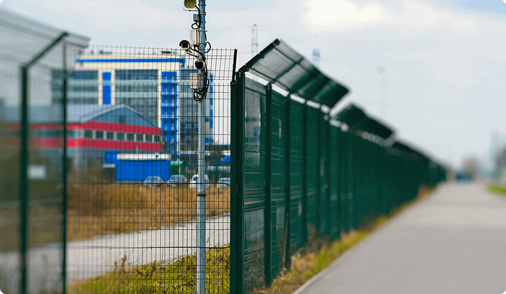 A long, green high-security fence with mounted surveillance cameras, protecting an industrial perimeter