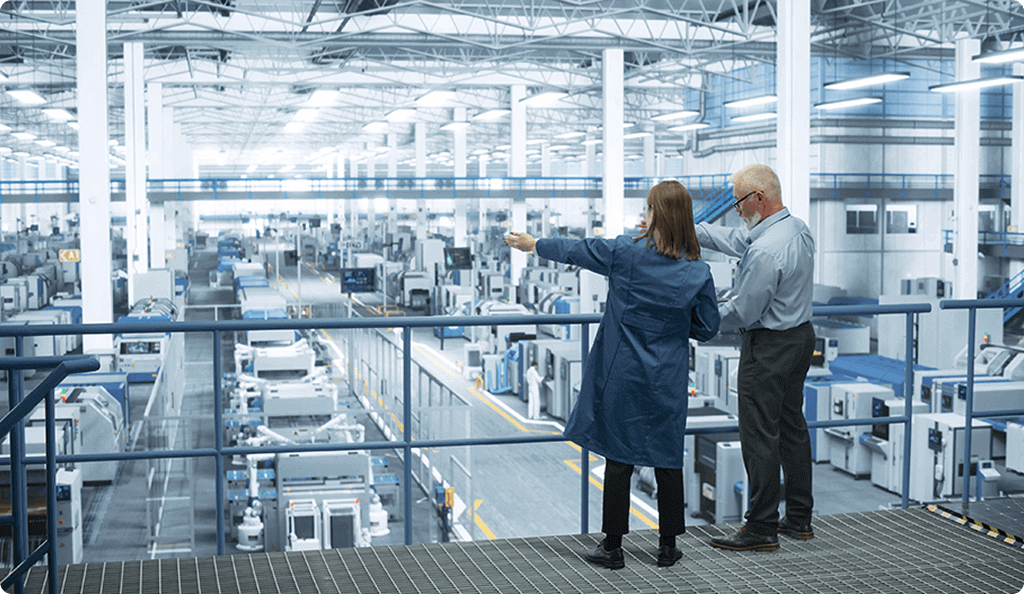 Two managers on an elevated walkway overlooking the floor of a large, modern manufacturing facility