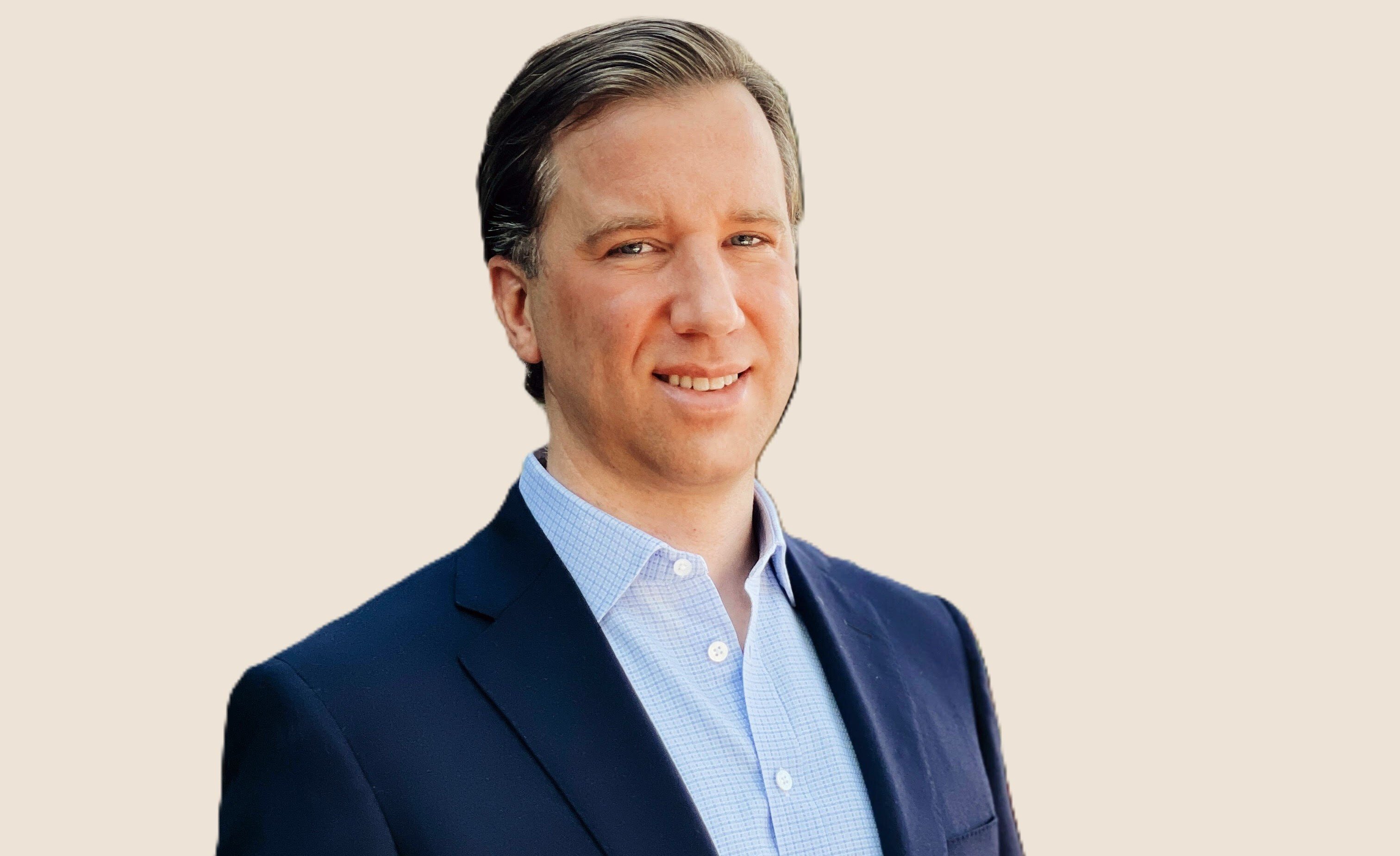 A professional headshot of a smiling man with a beard and glasses, wearing a blue shirt and a blue jacket